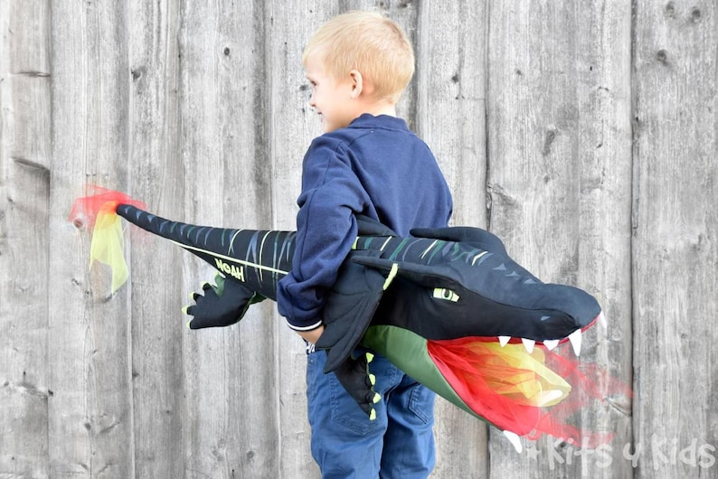 May include: A child wearing a blue shirt holds a black and green dragon puppet with a red and yellow fire mouth. The dragon has a green and yellow stripe pattern and the text "Woah" is visible on the side.