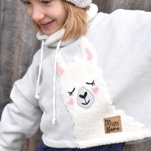 Puede incluir: Una sudadera con capucha gris claro con una aplicación de llama blanca y esponjosa. La llama tiene orejas rosas y los ojos cerrados. La sudadera tiene un parche marrón con el texto "No prob llama" en la parte inferior derecha.