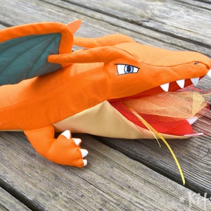 May include: Orange and green plush dragon toy with a yellow ribbon hanging from its mouth. The dragon's mouth is open and has a red and orange tulle inside.