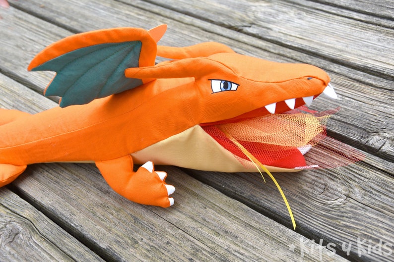 May include: Orange and green plush dragon toy with a yellow ribbon hanging from its mouth. The dragon's mouth is open and has a red and orange tulle inside.