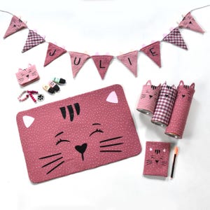 Könnte beinhalten: Eine Sammlung von altrosa Katzen-Artikeln, bestehend aus einer Wimpelkette mit der Aufschrift JULIE, einer rechteckigen Matte, drei zylindrischen Behältern, einem kleinen Beutel, einem Notizbuch, einem Perlenarmband und drei Stiften mit weißen Punkten.
