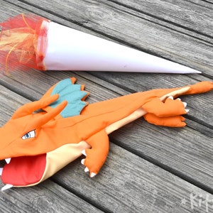 May include: Orange and teal dragon puppet with an open mouth and white teeth. The puppet is lying on a wooden surface with a white cone and red and yellow tulle attached to the end of the puppet's tail.