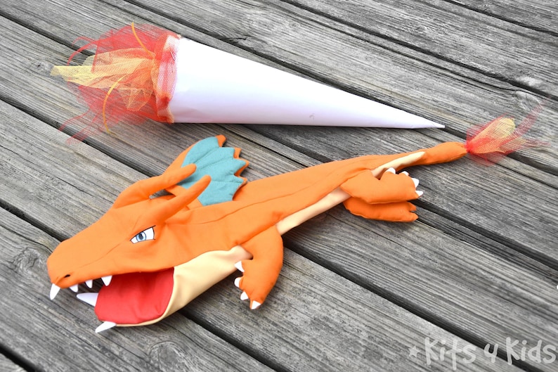 May include: Orange and teal dragon puppet with an open mouth and white teeth. The puppet is lying on a wooden surface with a white cone and red and yellow tulle attached to the end of the puppet's tail.