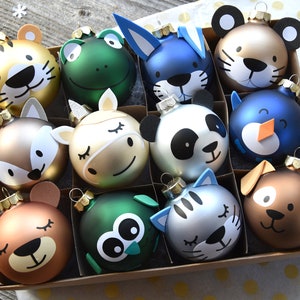 Può includere: Una scatola di 12 addobbi natalizi con diversi disegni di animali. Gli addobbi sono fatti di vetro e sono dipinti in vari colori, tra cui oro, argento, verde, blu e marrone. Ogni addobbo ha un viso di animale diverso, come una tigre, una rana, un coniglio, un panda, un pinguino, un gatto e un cane.