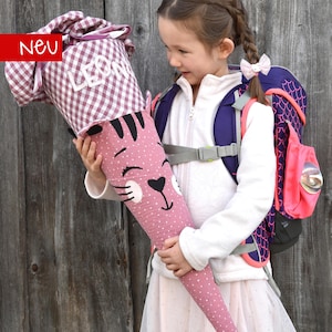 May include: A pink cat-themed school cone with white polka dots and black stripes, held by a child. The top is checkered with the name "LEON". A red banner with "Neu" is in the corner. A purple and pink backpack is also visible.