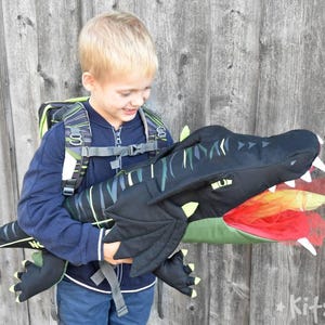 May include: A child wearing a black and green backpack with a dragon costume attached. The dragon has a black head with a green mouth and red and orange flames coming out of its mouth. The child is smiling and looking at the dragon.
