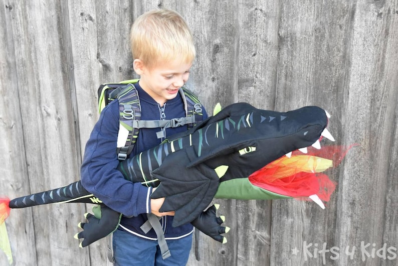 May include: A child wearing a black and green backpack with a dragon costume attached. The dragon has a black head with a green mouth and red and orange flames coming out of its mouth. The child is smiling and looking at the dragon.