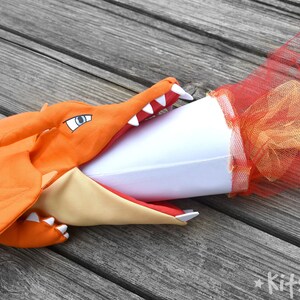 May include: Orange dragon puppet with a white cone in its mouth. The cone is decorated with red and gold tulle.