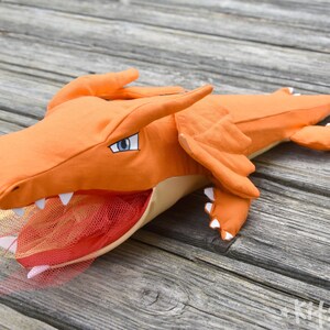 May include: Orange and beige plush dragon toy with a red and orange tulle tail. The dragon has a large open mouth with white teeth and a blue eye.