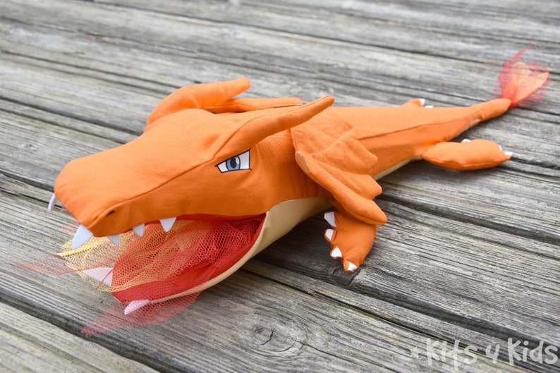 May include: Orange and beige plush dragon toy with a red and orange tulle tail. The dragon has a large open mouth with white teeth and a blue eye.