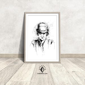 Può includere: Disegno a inchiostro nero e bianco di una persona con un cappello, incorniciato in una cornice di legno chiaro. L'opera d'arte presenta un ritratto dettagliato con linee ed elementi espressivi, creando un effetto strutturato. L'opera d'arte è appoggiata a un muro.