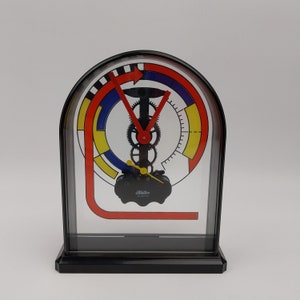 Puede incluir: Un reloj blanco y negro con una esfera roja, amarilla y azul. El reloj tiene un diseño único con una esfera circular y una flecha roja apuntando hacia la derecha. El reloj está hecho de plástico y tiene un movimiento de cuarzo. El reloj está etiquetado como "Mattel Quartz".