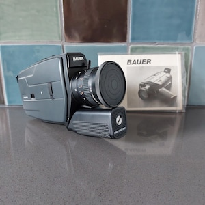 Op de afbeelding: Een grijze Bauer Super 8 filmcamera met een zwarte lens en handvat. De camera heeft de merknaam "Bauer" op de bovenkant gedrukt. Een handleiding met het Bauer-logo en een camera-illustratie staat op de achtergrond.