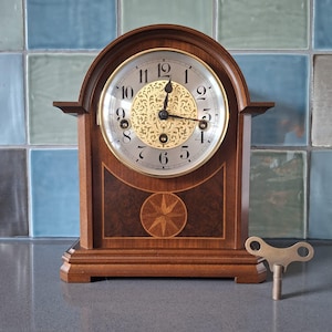 Op de afbeelding: Een houten mantelklok met een lichtgekleurde wijzerplaat en goudkleurige accenten. De klok heeft een decoratief sterrenpatroon op het onderste gedeelte en er ligt een sleutel naast. De klok is ongeveer 30 cm hoog.