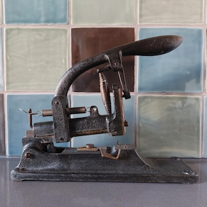 Puede incluir: Un punzón de papel antiguo de hierro fundido con un acabado oscuro y desgastado. La herramienta presenta un mango curvo, un mecanismo de resorte y una base con una matriz de corte. El fondo es una pared de azulejos con tonos azules, verdes y marrones.