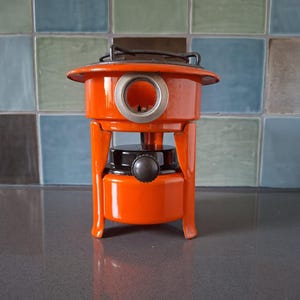 Op de afbeelding: Een feloranje draagbaar kampeerfornuis met een zwart branderrooster aan de bovenkant. Het fornuis is voorzien van een rond kijkvenster met een zilveren rand, een zwarte brandstoftankdop en drie stevige oranje poten onder het lichaam.