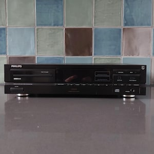 Puede incluir: Un reproductor de CD Philips negro con una placa frontal plateada y una variedad de botones y controles. El reproductor está en una encimera gris.