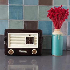 Könnte beinhalten: Ein vintage braunes Bakelit-Radio mit weißem Zifferblatt und verchromten Knöpfen. Das Radio steht auf einer Arbeitsplatte mit einer türkisfarbenen Vase, die mit roten Blumen gefüllt ist.