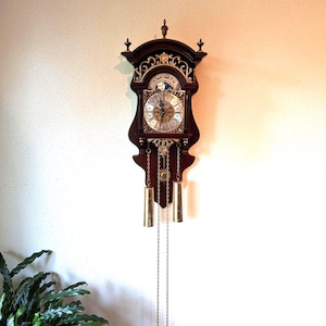 May include: A dark wood wall clock featuring a moon phase dial, gold-toned filigree accents, and a white clock face with black Roman numerals. Two brass-colored cylindrical weights hang from chains below the clock body, which is topped with a finial.