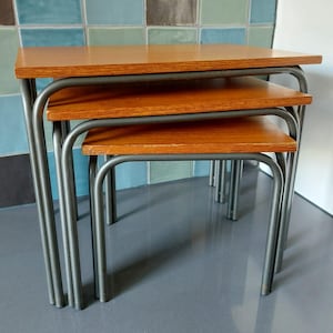 Puede incluir: Un conjunto de tres mesas nido con tapa de madera y marco de metal plateado. Las mesas están apiladas una encima de la otra.