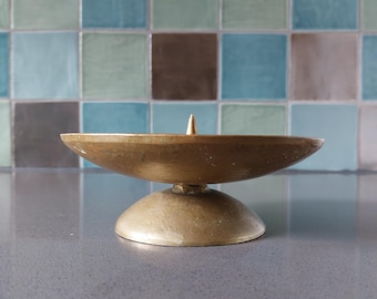 May include: A vintage brass candle holder with a shallow, bowl-shaped top and a small, conical spike in the center. The candle holder sits on a rounded base. The background features a tiled wall with shades of blue, green, and brown.