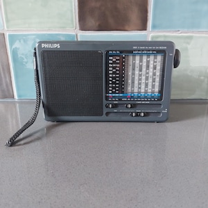 Könnte beinhalten: Ein graues tragbares Philips-Radio mit schwarzem Lautsprechergrill und Drehknopf. Das Radio hat einen Trageriemen und ein Display mit Frequenzbändern. Der Text "PHILIPS" ist oben sichtbar.