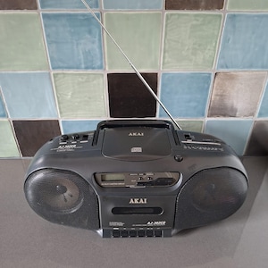 Op de afbeelding: Een donkergrijze, draagbare Akai AJ-302CD stereoboombox met een cd-speler aan de bovenkant, een cassettedeck aan de voorzijde, twee luidsprekers aan de zijkanten, een digitaal display en een lange, dunne, zilverkleurige telescopische antenne.
