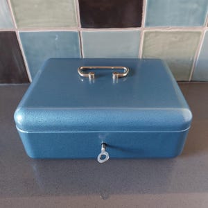 Könnte beinhalten: Eine blaue Metallkassette mit silberfarbenem Griff und Schlüsselloch. Die Box hat eine strukturierte Oberfläche und eine rechteckige Form. Der Griff ist gewölbt und an der Oberseite der Box befestigt. Das Schlüsselloch befindet sich vorne.
