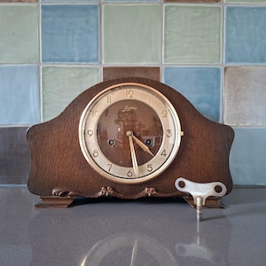 Puede incluir: Un reloj de sobremesa de madera marrón oscuro con parte superior arqueada y esfera circular dorada con números arábigos negros. El reloj tiene manecillas doradas y una llave de cuerda de metal plateado situada a la derecha de la base.