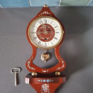 Peut inclure: Une horloge murale en bois rouge foncé avec un cadran circulaire, des chiffres romains et des détails floraux peints à la main. L'horloge comprend un pendule en laiton, une clé de remontage et une petite étagère à la base.
