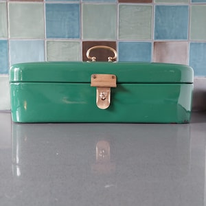 May include: A rectangular, emerald green metal box with a gold-colored handle and clasp. The box has rounded edges and a glossy finish. The handle is arched, and the clasp is rectangular with a keyhole. The box is sitting on a gray surface.