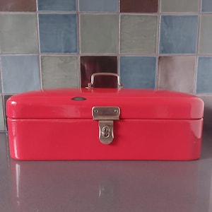 May include: A bright red metal bread box with a silver handle and clasp. The rectangular box has rounded edges and a small, circular emblem on the front. The background features a tiled wall with various shades of blue and green.
