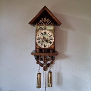 Peut inclure: Une horloge murale en bois avec un toit en pignon, dotée d'un cadran circulaire à chiffres romains et d'un paysage peint. L'horloge comprend une crête décorative en laiton, deux poids cylindriques en laiton suspendus et des chaînes en métal.
