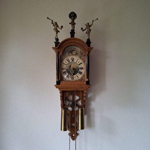 May include: A wooden wall clock featuring a central dial with Roman numerals and a nautical scene. The clock is topped with three brass figures, including two trumpeters and a central globe. Two brass weights hang from chains below the clock face.