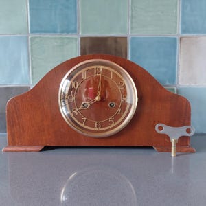 May include: A vintage wooden mantel clock with a clear glass face and gold-colored numerals. The clock has a key and a winding mechanism. The clock's face shows the time as approximately 12:00. The clock is set on a gray surface.