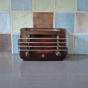Könnte beinhalten: Ein dunkelbrauner Vintage-Toaster in abgerundeter rechteckiger Form mit horizontalen Lamellen, zwei runden Drehknöpfen an der Vorderseite und einem kleinen Philips-Logo-Emblem in der Mitte unten. Das Gerät hat eine glänzende Oberfläche.