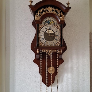 Peut inclure: Une horloge murale en bois foncé avec un cadran doré à chiffres romains, un indicateur de phases de lune et des ornements en filigrane doré. Trois chaînes pendent de la base, chacune portant un poids en laiton, avec un pendule en soleil.