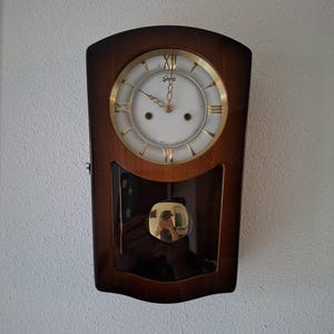 May include: A wall clock with a dark brown wooden frame and a white circular face featuring gold Roman numerals. The clock has a gold-coloured pendulum visible through a glass panel at the bottom. The brand name 'Gerses' is printed on the clock face.