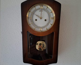 Op de afbeelding: Een wandklok met een donkerbruine houten lijst en een witte ronde wijzerplaat met gouden Romeinse cijfers. De klok heeft een goudkleurige slinger die zichtbaar is door een glazen paneel aan de onderkant. De merknaam 'Gerses' staat op de wijzerplaat.