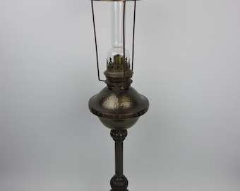 May include: An antique oil lamp with a clear glass chimney and a bronze-colored metal base. The lamp has a decorative, hammered texture on the central globe and a tall, slender stem. The lamp is designed to stand upright.