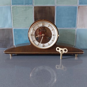 May include: A vintage wooden mantel clock with a round face and gold-tone detailing. The clock face is white with black numerals and hands. A gold-tone key is inserted. The clock sits on a dark surface. The clock measures approximately 20 cm in width.