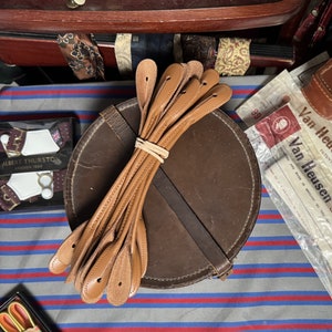 Peut inclure: Un lot de huit lanières en cuir marron avec des détails de couture et un trou à l'extrémité de chaque lanière. Les lanières sont attachées ensemble avec un élastique et sont posées sur un coffret rond en cuir marron.
