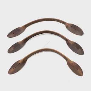 Peut inclure: Trois poignées en cuir marron avec un design incurvé. Chaque poignée a une extrémité ovale avec deux petits trous et une couture. Les poignées sont probablement pour un sac ou un bagage.