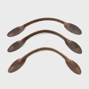 Peut inclure: Trois poignées en cuir marron avec un design incurvé. Chaque poignée a une extrémité ovale avec deux petits trous pour la fixation. Les poignées sont probablement destinées à un sac ou à un article similaire.