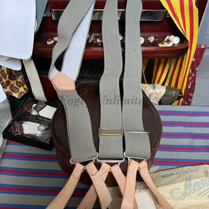 Op de afbeelding: Drie paar grijze bretels met leren banden en metalen clips. De bretels hangen aan een houten plank met andere accessoires.