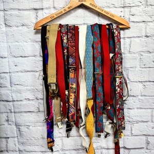 Puede incluir: Una colección de tirantes coloridos colgados de una percha de madera. La percha tiene el texto "The Four Feathers & Strandley", "33A Dover St. W.I." y "Phone Hyde Park 4591" impreso en ella.