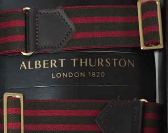 May include: A pair of suspenders with a striped pattern in dark green and red. The suspenders have brown leather accents and gold-colored metal clasps. The brand name "ALBERT THURSTON LONDON 1820" is printed on the box.