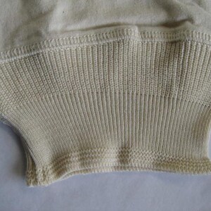 Könnte beinhalten: Nahaufnahme eines cremefarbenen Strickkleidungsstücks. Das gerippte Strickbündchen ist leicht ausgestellt und weist ein detailliertes, strukturiertes Muster auf. Der Stoff wirkt weich und hat eine subtile, natürliche Farbe.