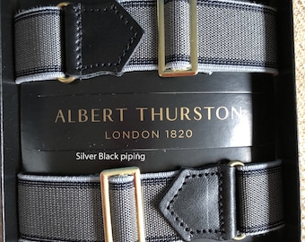 May include: A pair of gray and black suspenders with silver and black piping. The suspenders have a gold buckle and are labeled "Albert Thurston London 1820".