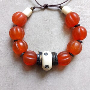 May include: A handmade bracelet featuring orange, ribbed beads interspersed with black spacers and cream-colored, cylindrical beads. The bracelet is strung on a brown cord, tied at the top. The beads have a translucent quality.